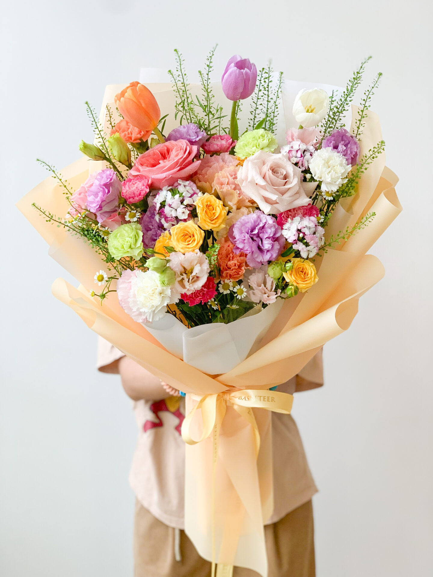 A person holds a large, vibrant bouquet of flowers wrapped in light yellow paper and secured with a yellow ribbon. The bouquet features a variety of blooms, including pink and purple roses, orange tulips, white carnations, and other colorful flowers, against a plain white background.