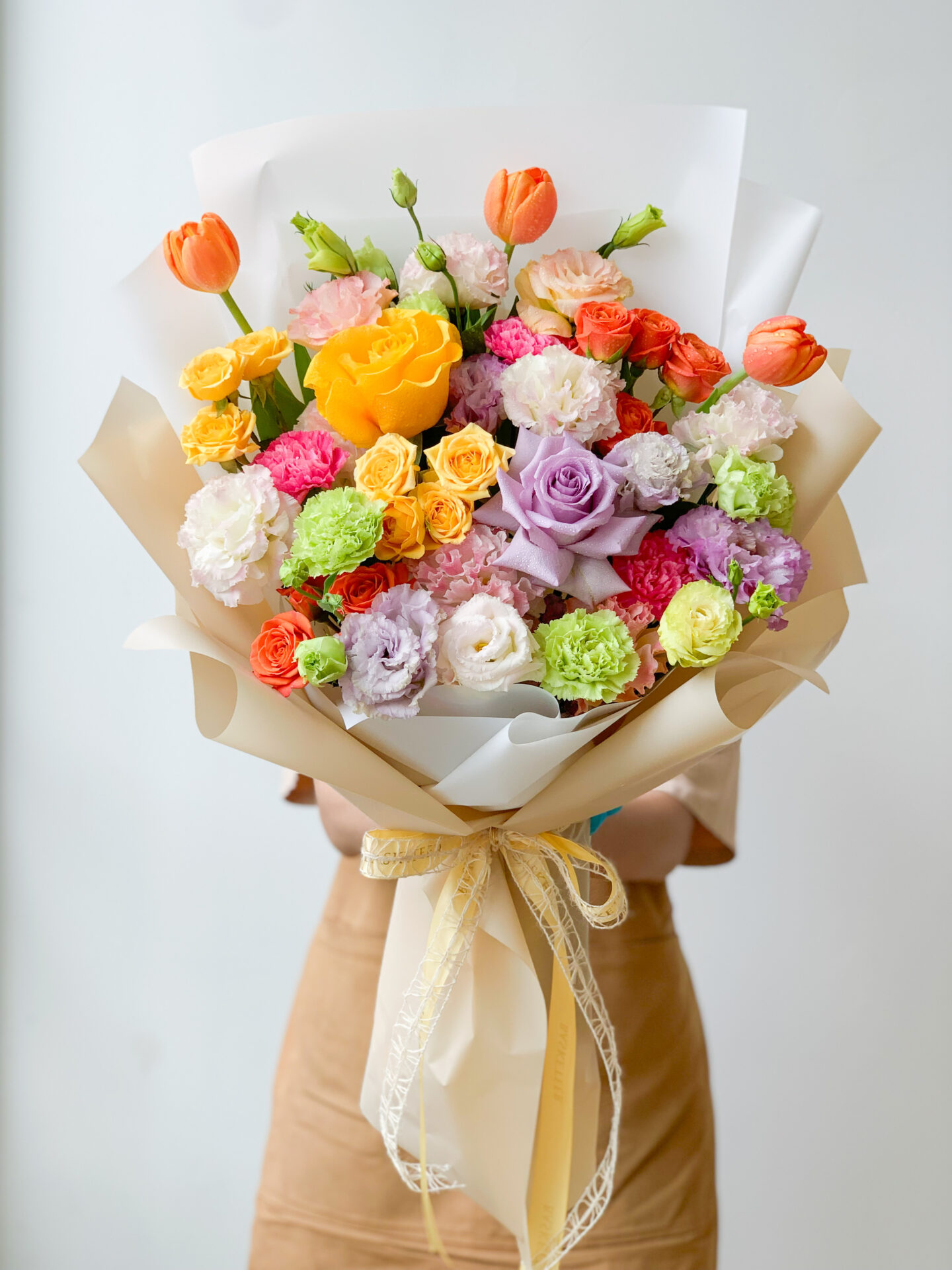 A person in a beige dress holding a vibrant bouquet of flowers wrapped in white paper and tied with a ribbon. The bouquet includes orange tulips, yellow roses, purple and white roses, and various colorful blossoms against a plain background.