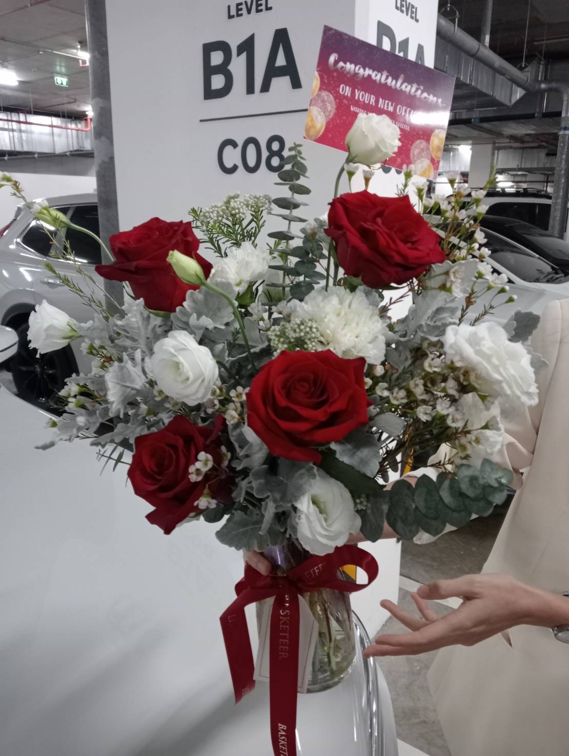 A Rustic Charm Rose In Vase arrangement of red and white roses with greenery, topped with a “Congratulations on your new car” card, sits on a white car in an indoor car park. A person's hand is visible beside the bouquet.