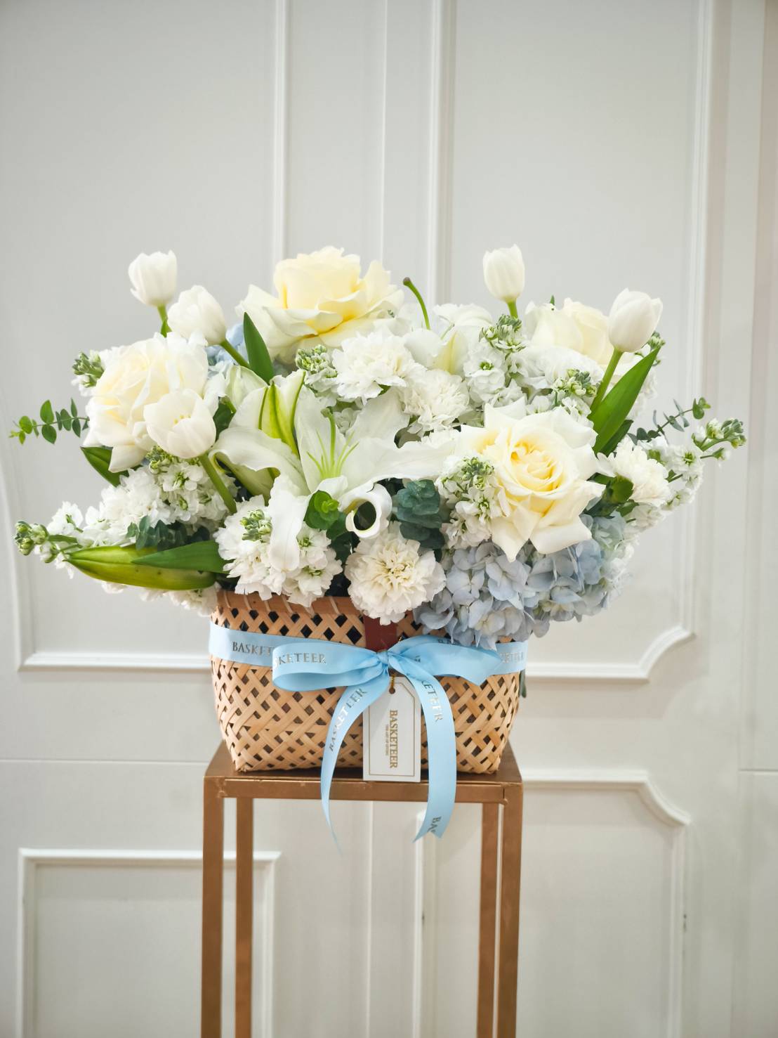 Elegant woven basket with white roses, tulips, lilies, carnations, and pastel blue hydrangeas, accented with a blue Basketeer ribbon.