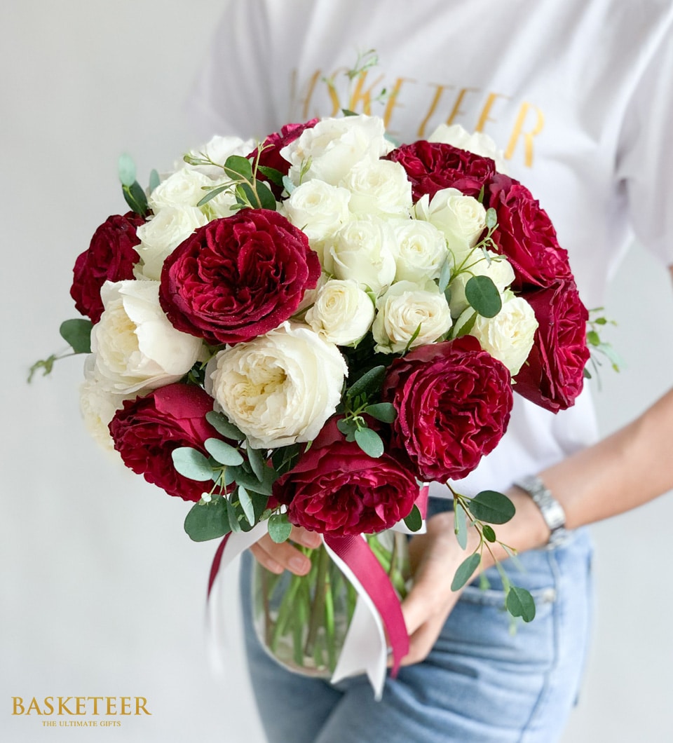 A person elegantly holds an opulent vase brimming with a lavish bouquet of red and white roses, meticulously accented with lush green foliage. The individual is adorned in a pristine white shirt, subtly showcasing the prestigious 