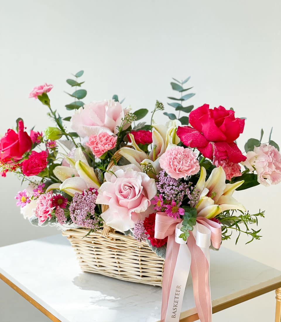 An opulent wicker basket overflowing with a vibrant selection of flowers, featuring pink and red roses, elegant pink carnations, pristine white lilies, and rich greenery. The luxurious arrangement rests on a pristine white surface. A delicate pink ribbon adorned with the Basketeer logo is gracefully attached to the handle, symbolizing the brand's commitment to sophistication and elegance. The backdrop remains an immaculate white, enhancing the allure of this exquisite floral display by Basketeer.