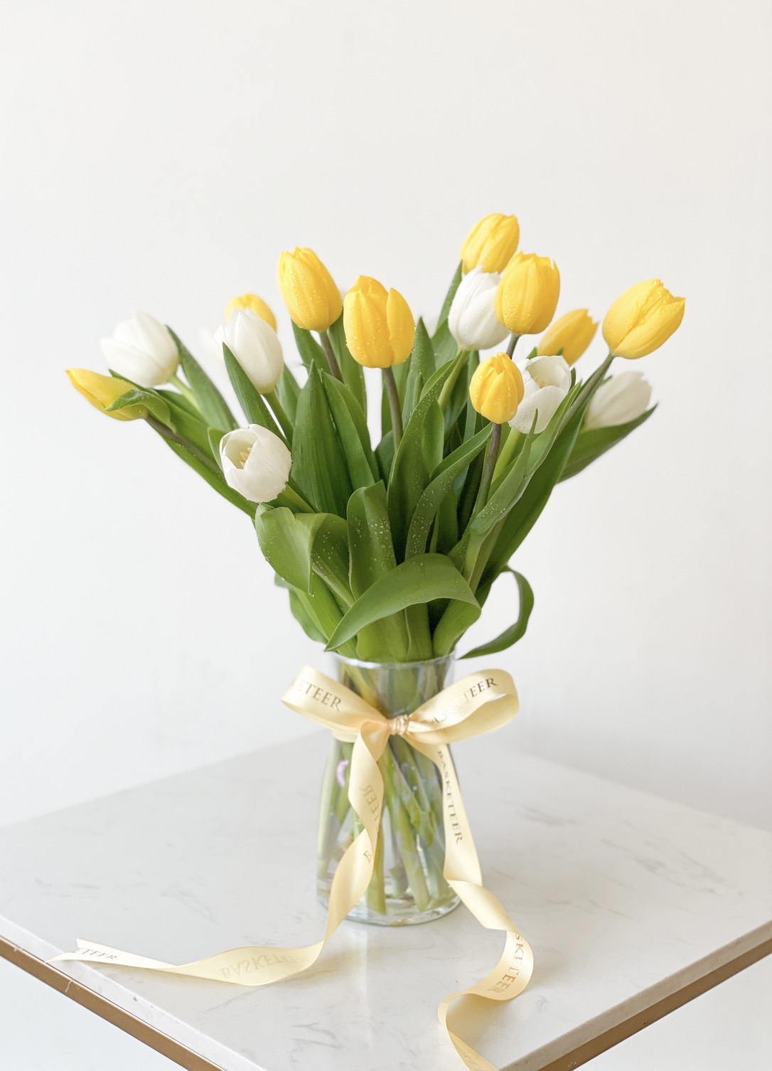 A fresh tulip arrangement with yellow and white tulips in a glass vase, tied with a golden satin ribbon.