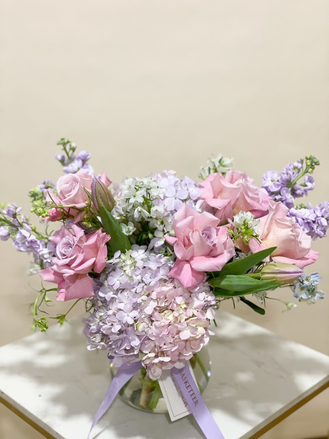 A luxurious pastel floral vase arrangement featuring soft pink roses, lilac hydrangeas, and delicate blossoms in a clear glass vase, wrapped with a silk ribbon.