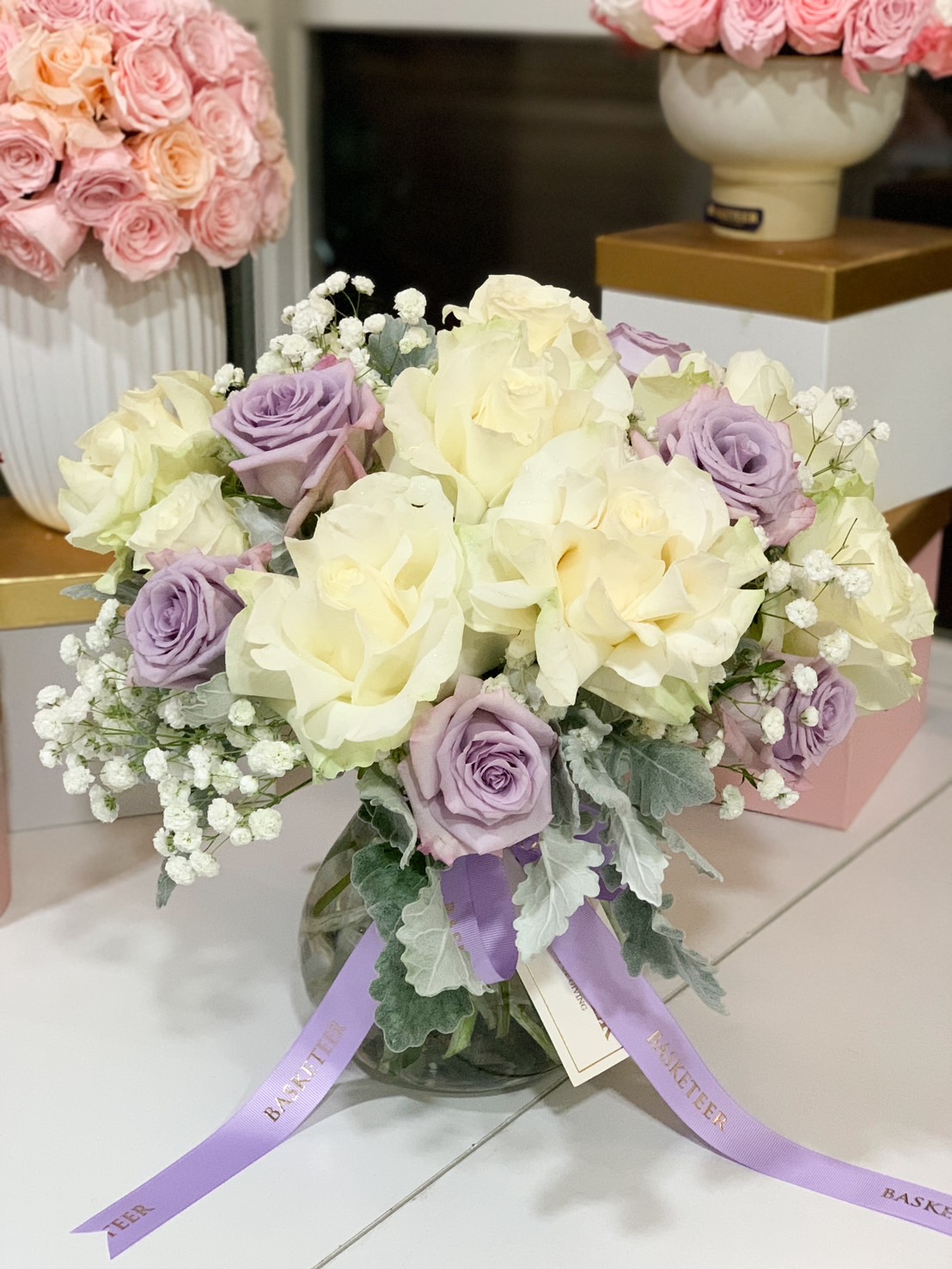 A luxurious white and lavender rose arrangement in a clear glass vase, adorned with baby’s breath and lush greenery, tied with a lavender satin ribbon.