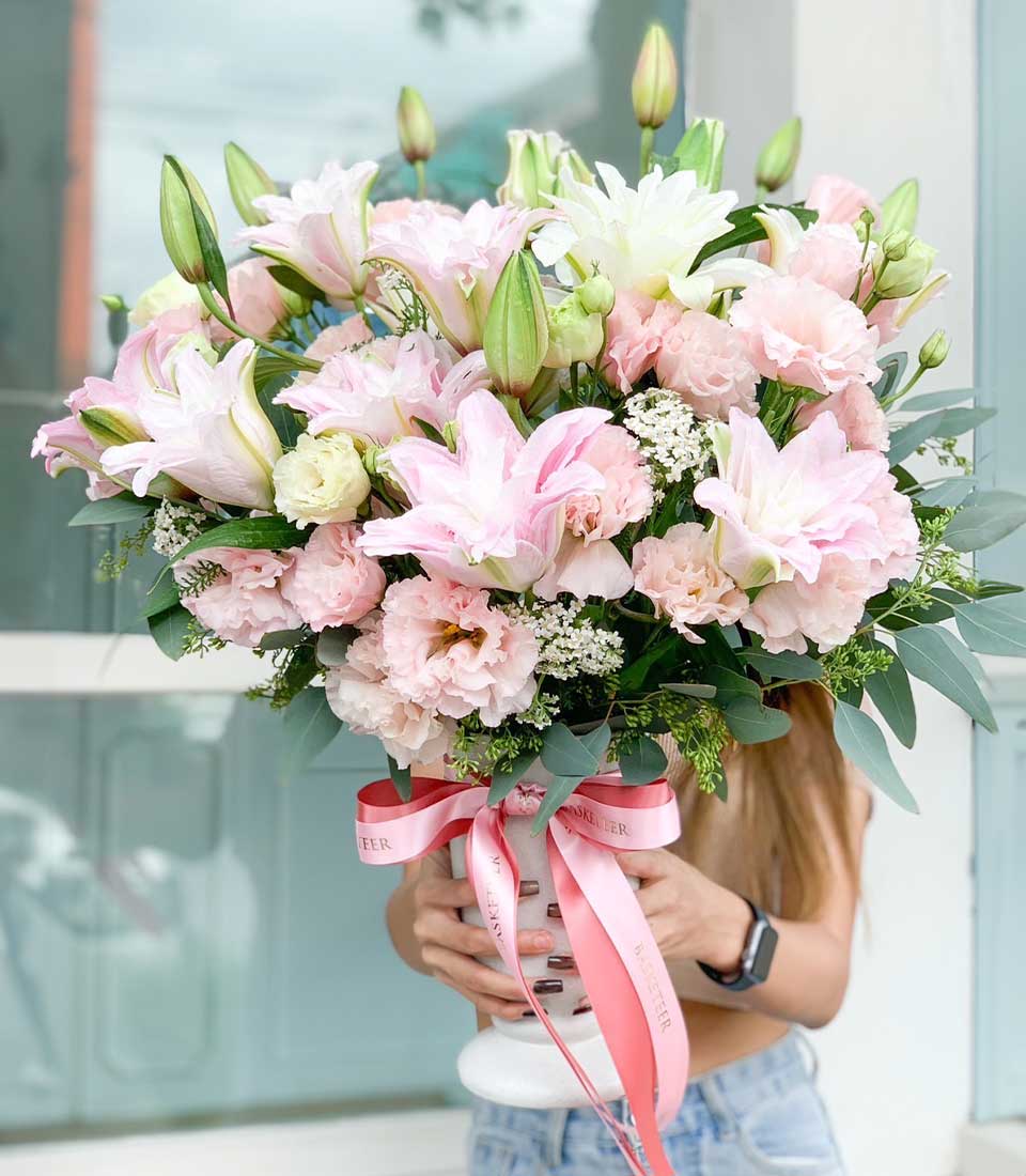A person holding a large bouquet of pink and white flowers, including lilies and carnations, wrapped in pink ribbon. The background is a light-colored wall.