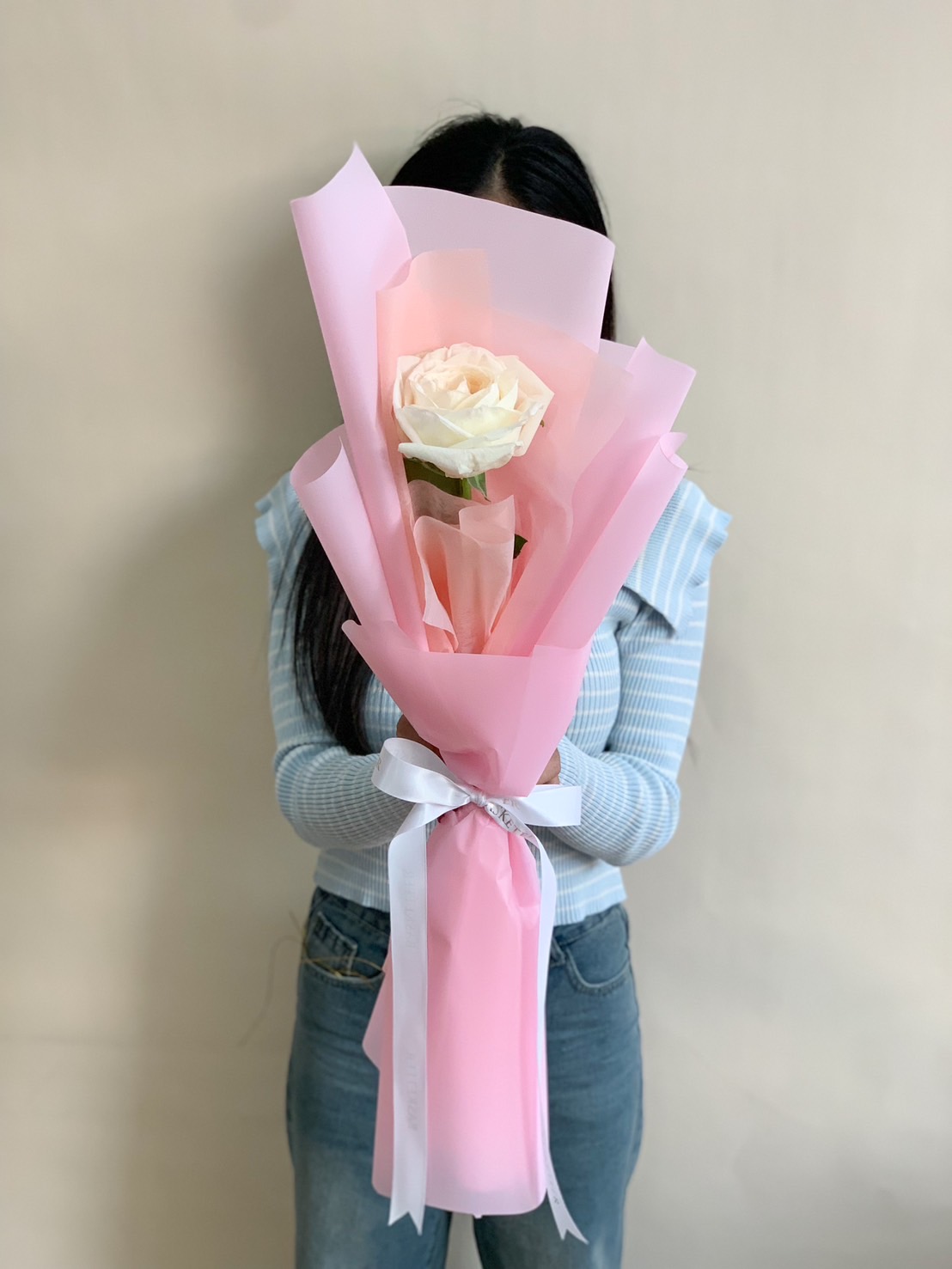 A beautiful single white rose wrapped in soft pink paper with a white ribbon, held by a person wearing a light blue sweater, creating a delicate and romantic aesthetic.