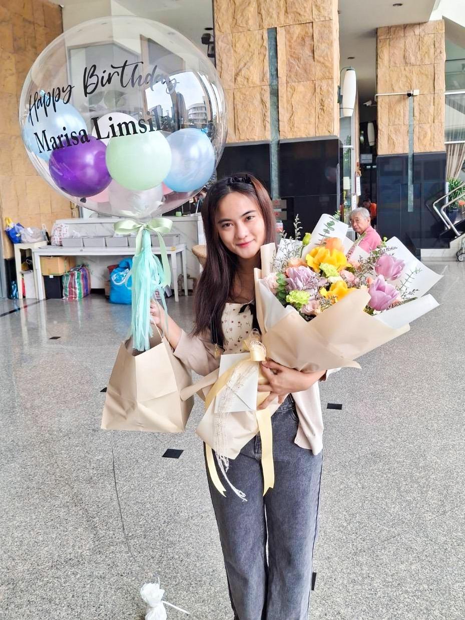 A woman stands indoors holding a bouquet of bright flowers and a balloon. The balloon is translucent with purple, green, and white smaller balloons inside, and it has "Happy Birthday Marisa Lims" written on it. She smiles and holds a paper bag containing a chocolate cake set in her other hand.