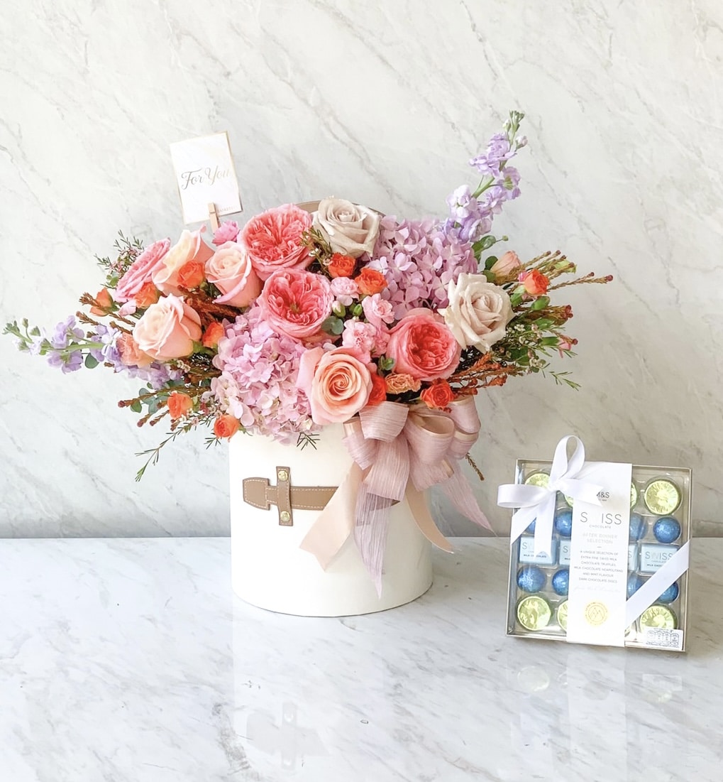 A white hatbox filled with a mix of pink roses, purple hydrangeas, and other colorful flowers, adorned with a pink ribbon and a small card. Next to it is a box of assorted chocolates wrapped with a white ribbon, placed on a marble surface.
