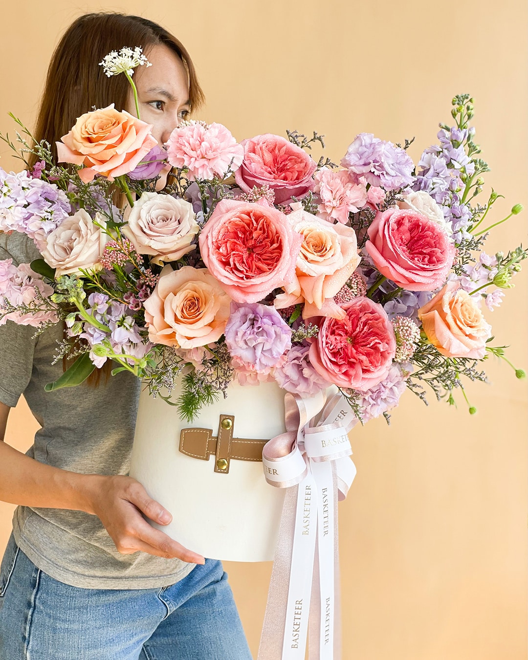 A person holding a large, round white vase filled with an arrangement of pink, peach, and lavender flowers. The vase has a tan strap attached, and a white ribbon with the word 