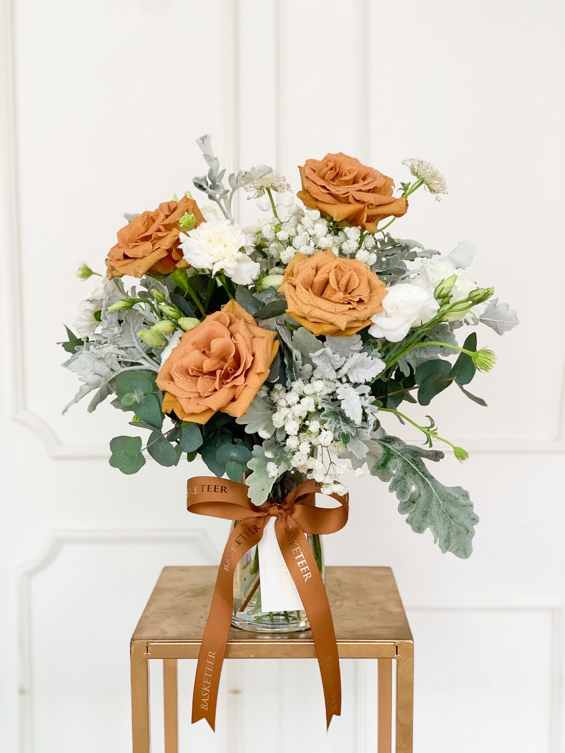 Earth-tone rose bouquet in a clear glass vase with white accent flowers and brown ribbon