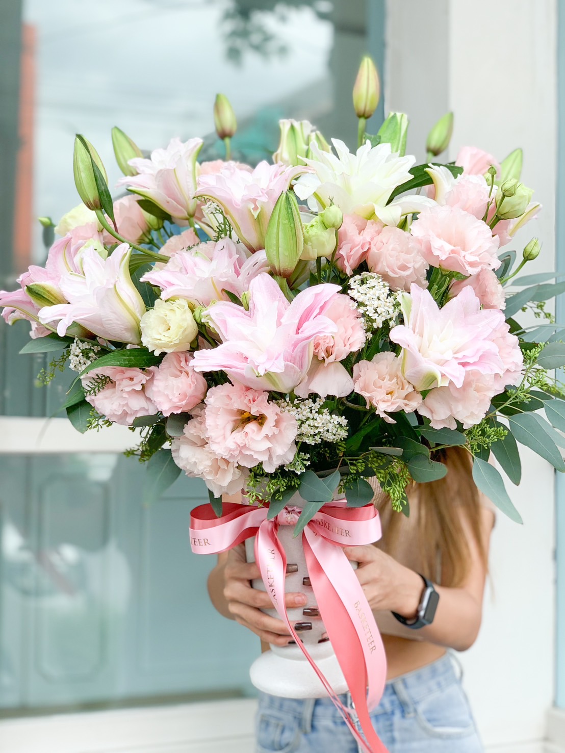 A person in a sleeveless top is elegantly holding an opulent bouquet of luxurious pink and white flowers, prominently featuring exquisite roses, lilies, and lush greenery. The bouquet is artfully tied with a silky pink ribbon embossed with the Basketeer logo, adding a touch of sophistication. The beautifully designed floral arrangement obscures the person's face, allowing the stunning blooms to take center stage. In the serene background, soft light filters through a window overlooking tranquil outdoor elements, enhancing the inviting ambiance.