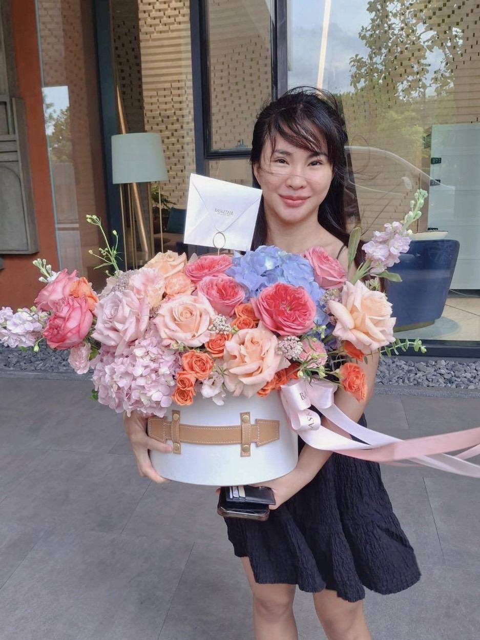 A person in a black dress holds a large, round white box filled with an assortment of vibrant flowers, including roses and hydrangeas, adorned with ribbon. The person is smiling and standing in front of a building with large windows and a modern design.