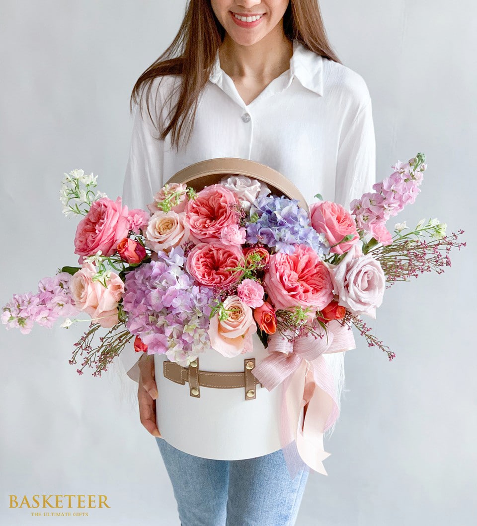 ผู้หญิงในชุดเสื้อสีขาวถือกล่องหมวกทรงกลมสีขาวที่เต็มไปด้วยดอกไม้สีชมพู สีพีช และสีม่วงหลากสีสัน กล่องนี้มีเข็มขัดหนังสองเส้น และการจัดประกอบด้วยดอกกุหลาบและดอกไม้นานาชนิดอื่นๆ โลโก้สำหรับ 