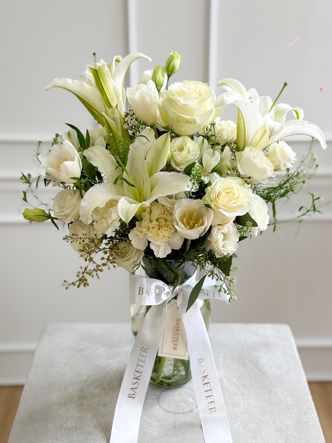 A Pure White Vase Arrangement filled with a bouquet of white lilies, roses, and assorted greenery, adorned with white ribbons labelled 