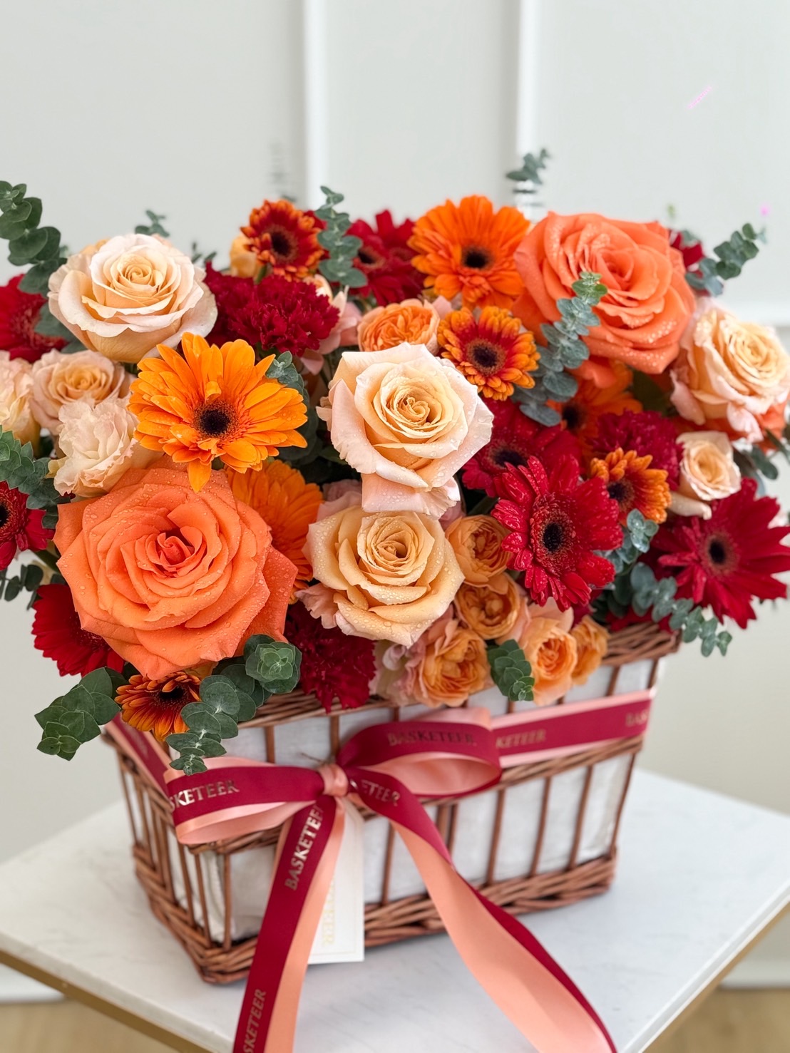 Luxury basket of orange and red roses with bright gerberas and greenery, tied with red satin ribbons.