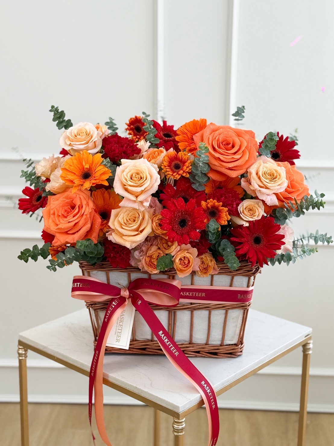 Elegant basket of orange roses, peach roses, and red gerberas with eucalyptus greenery, tied with dual satin ribbons.