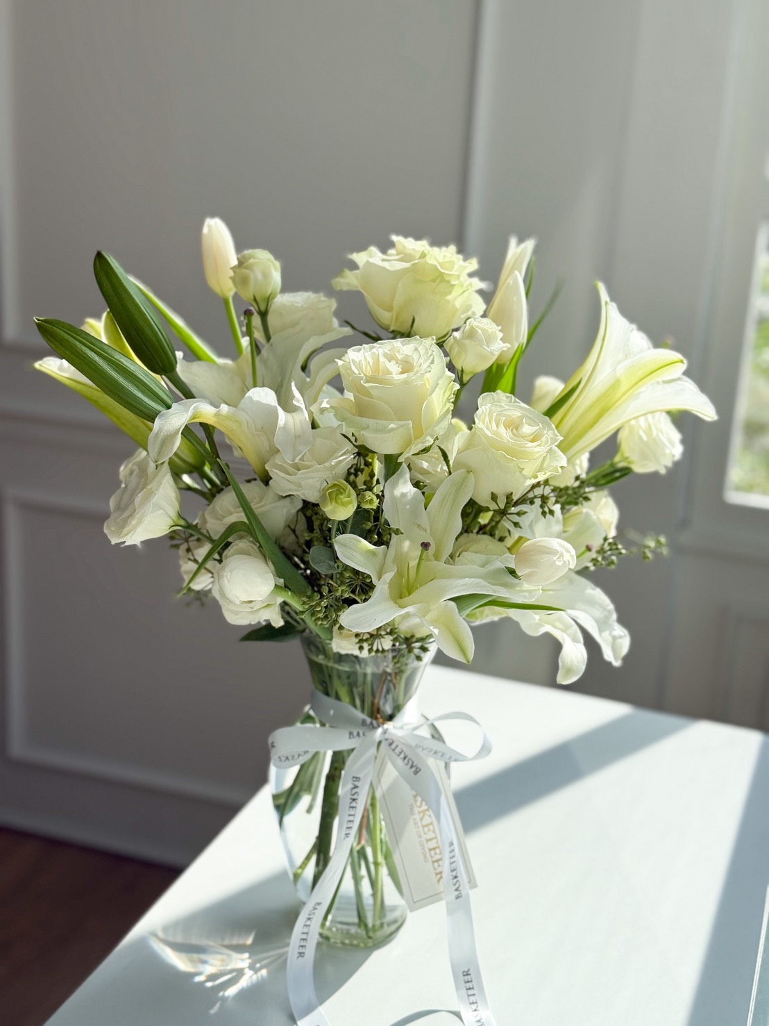 A Pure White Vase Arrangement of roses, lilies, and tulips sits on a light-coloured table in sunlight. The clear glass vase is tied with a white ribbon printed with 