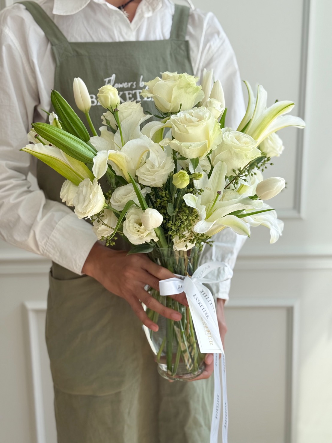 A person wearing a white shirt and green apron holds a Pure White Vase Arrangement filled with white roses, lilies, and greenery, tied with a white ribbon, standing against a light-coloured background.