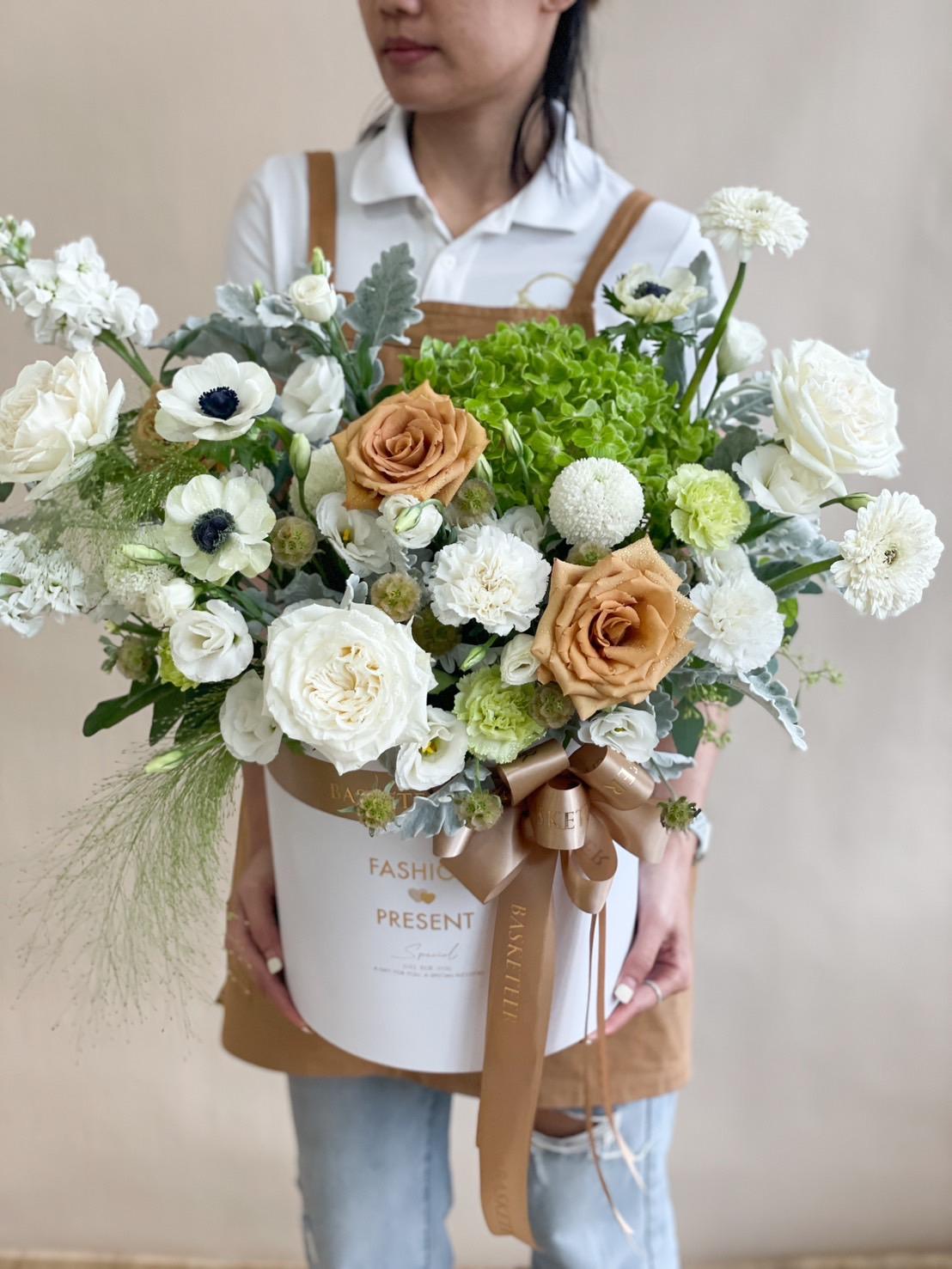 A person holding a large, elegantly arranged flower bouquet in a white hat box. The bouquet features white and beige roses, green hydrangeas, and various white flowers. The person is wearing a white shirt and brown apron, and their face is partially obscured.