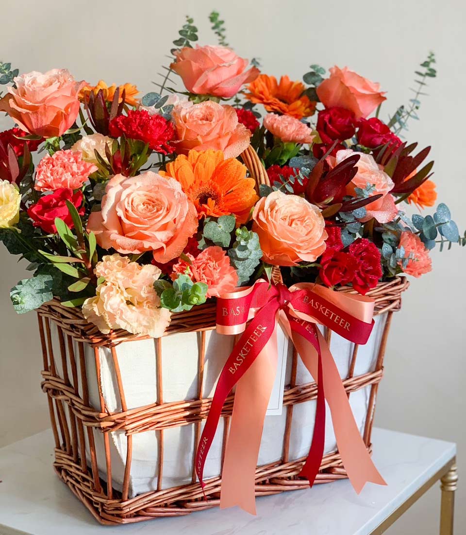 Floral basket with vibrant mixed blooms including roses, gerberas, and carnations, arranged in a woven wicker basket with a red ribbon.