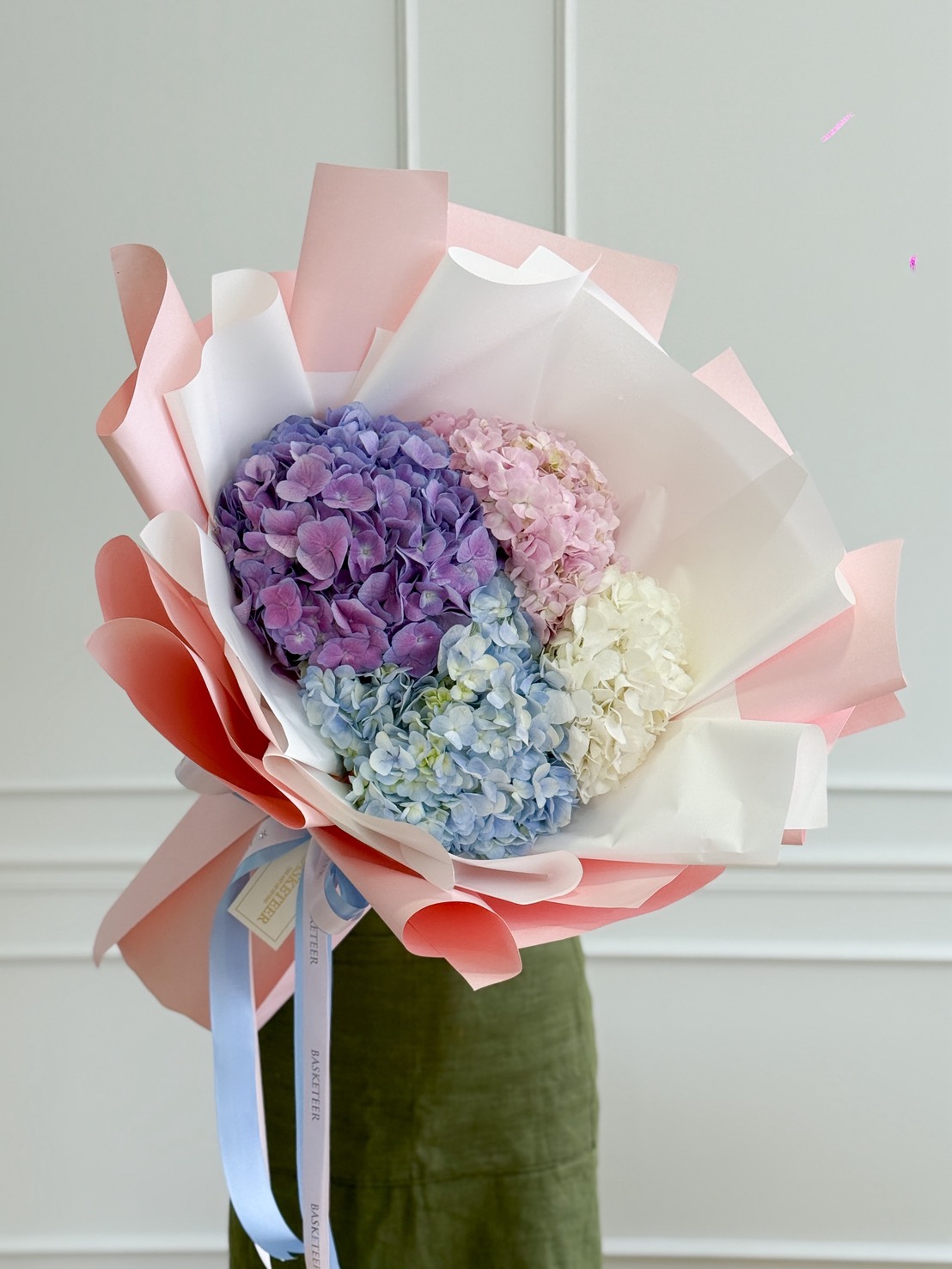A Pastel Hydrangeas Bouquet in shades of purple, pink, blue, and white is wrapped in pink and white paper with a blue ribbon, held in front of a white wall.