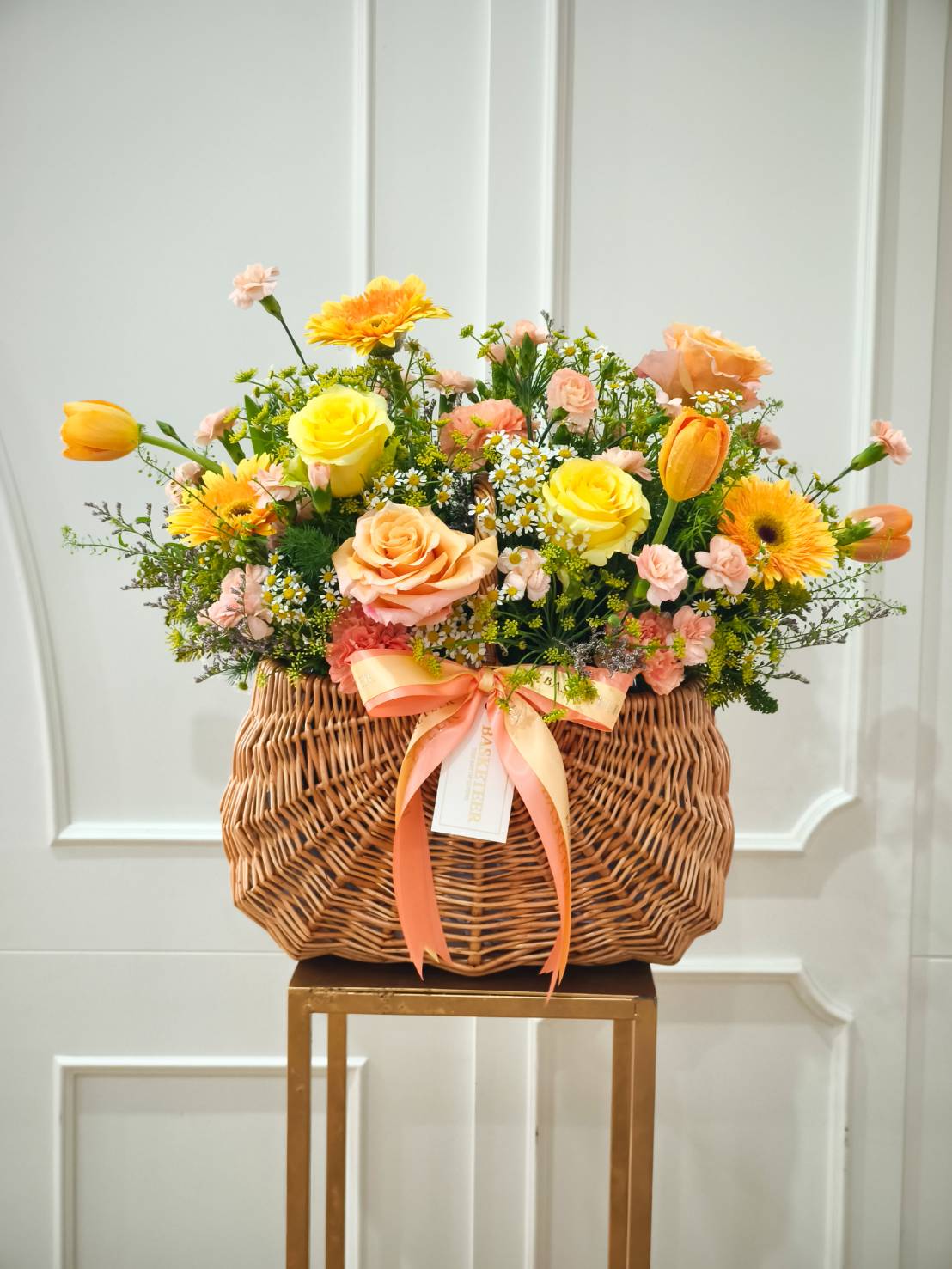 Woven basket filled with yellow roses, peach roses, tulips, gerberas, chamomile, and greenery, tied with a peach satin ribbon.
