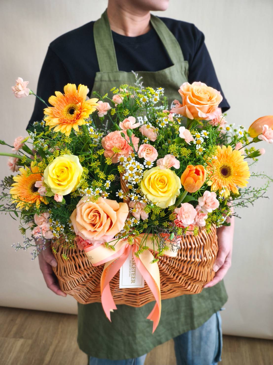 Woven basket with yellow roses, peach roses, orange tulips, yellow gerberas, chamomile, and peach spray carnations, tied with satin ribbon.