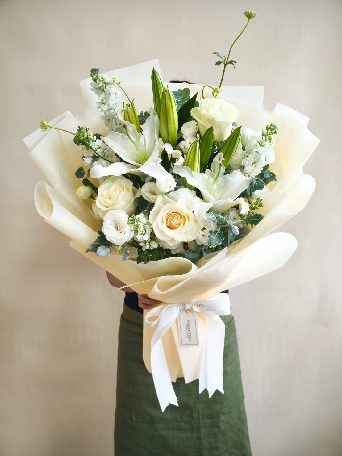 Large bouquet of white lilies, white roses, and seasonal white blooms wrapped in ivory paper with a satin ribbon.