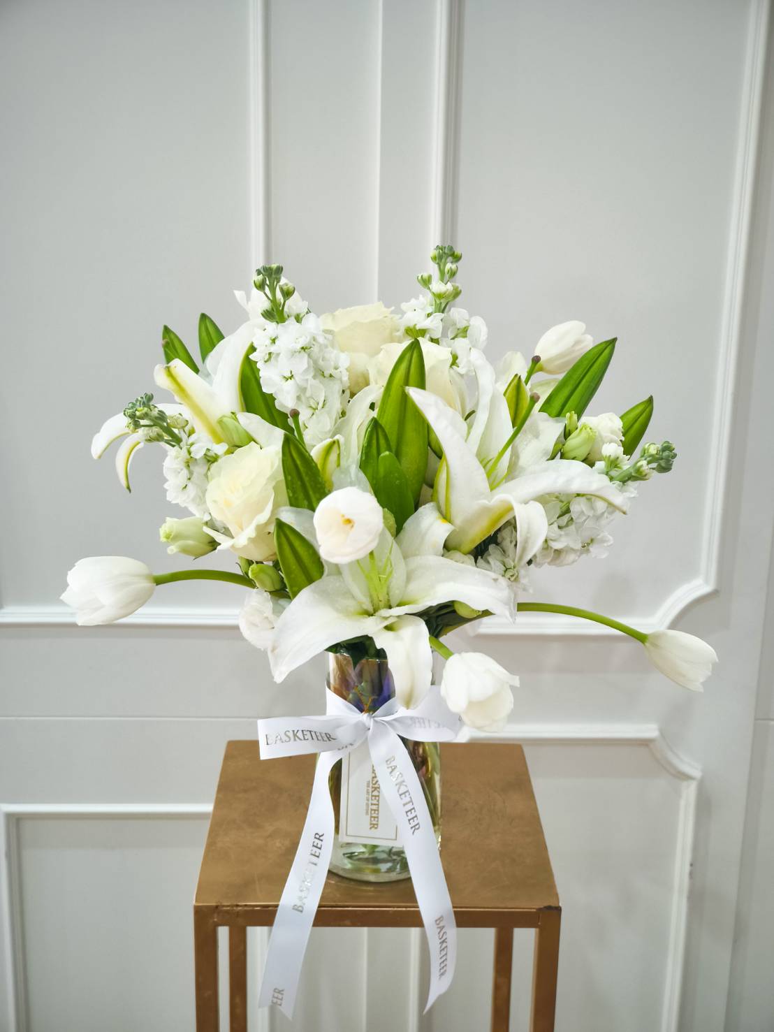 Glass vase filled with fresh white lilies, tulips, roses, and seasonal blooms, tied with a white satin ribbon.