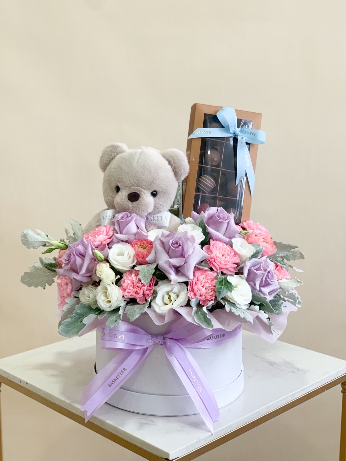 Flower gift box with pink and purple roses, teddy bear, and premium chocolates in elegant round box.