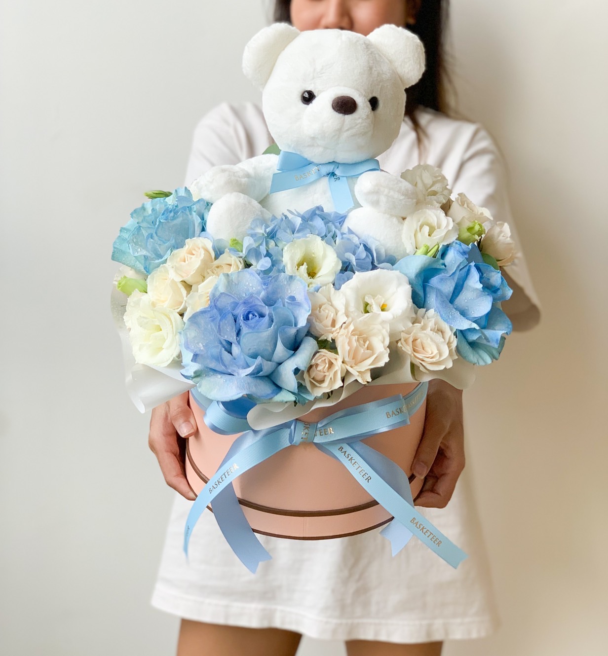 A person in a white dress holds a pink hatbox filled with blue and white flowers, topped with a white teddy bear. The arrangement includes ribbons tied around the box.