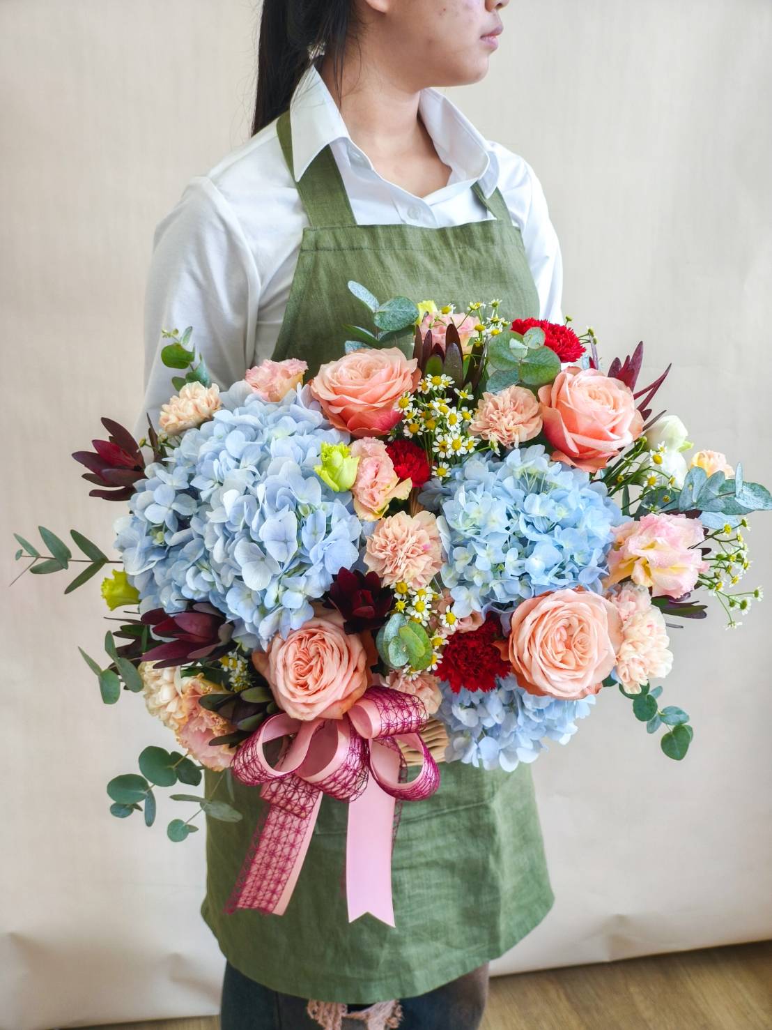 Large bouquet with blue hydrangeas, peach roses, carnations, and red accents, decorated with greenery and a pink ribbon.