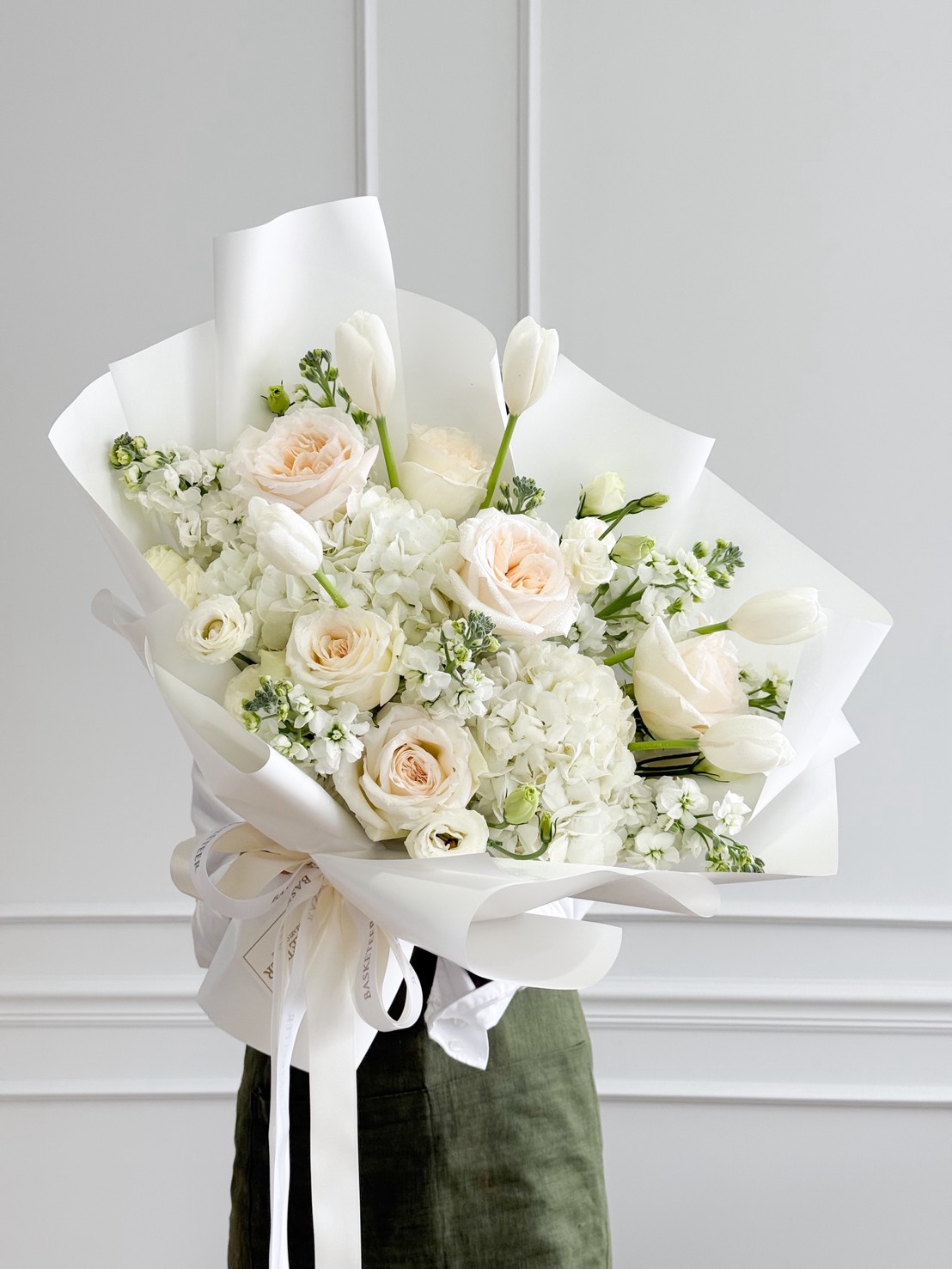 A person holds a Luminous White Roses Bouquet, featuring white roses, tulips, and hydrangeas wrapped in white paper with a ribbon. The background displays a white wall adorned with elegant decorative moulding.