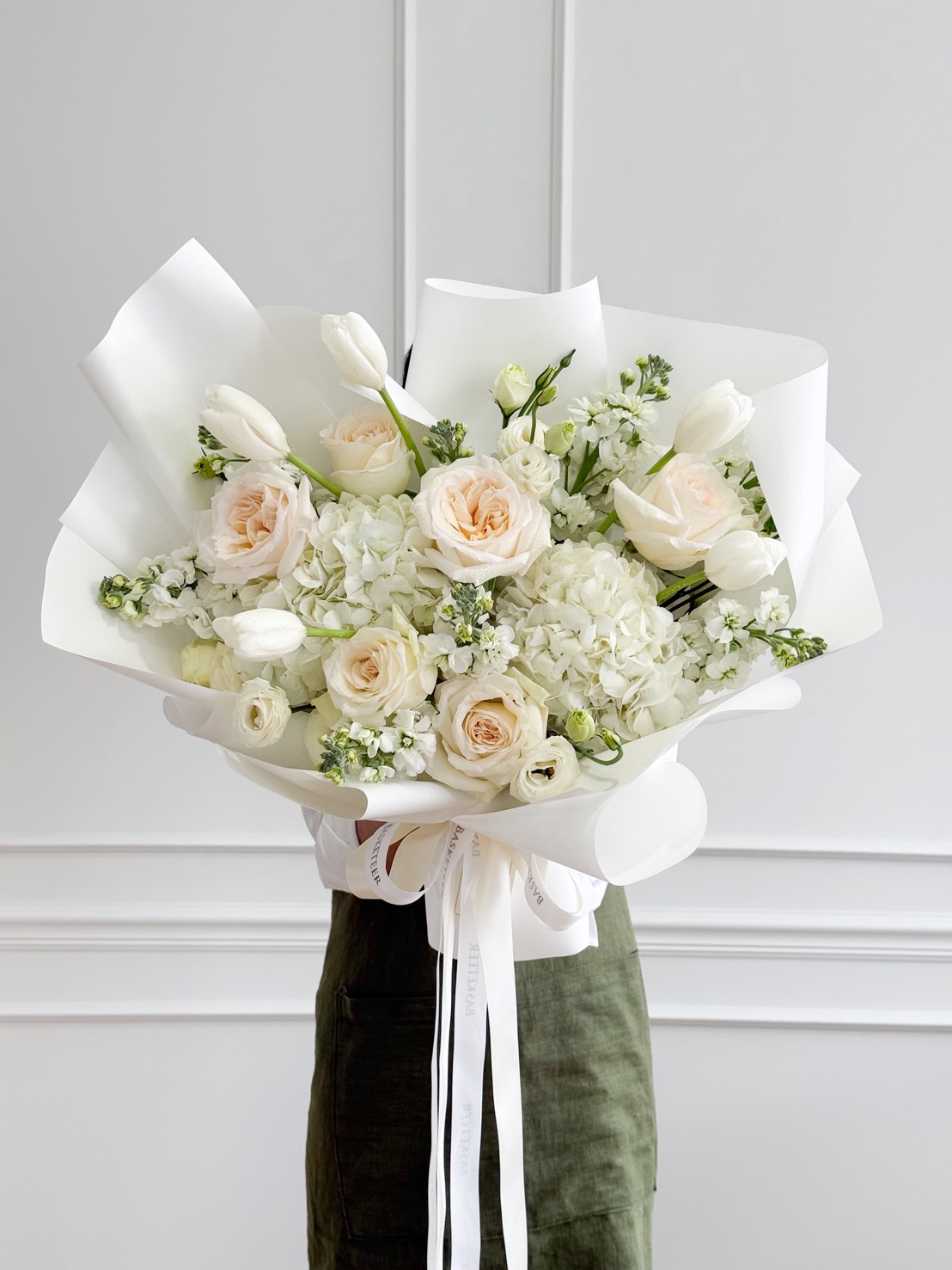 A person in a green outfit holds a Luminous White Roses Bouquet, featuring white and pale peach flowers like hydrangeas and tulips, beautifully wrapped in white paper and tied with ribbon, set against a white wall background.