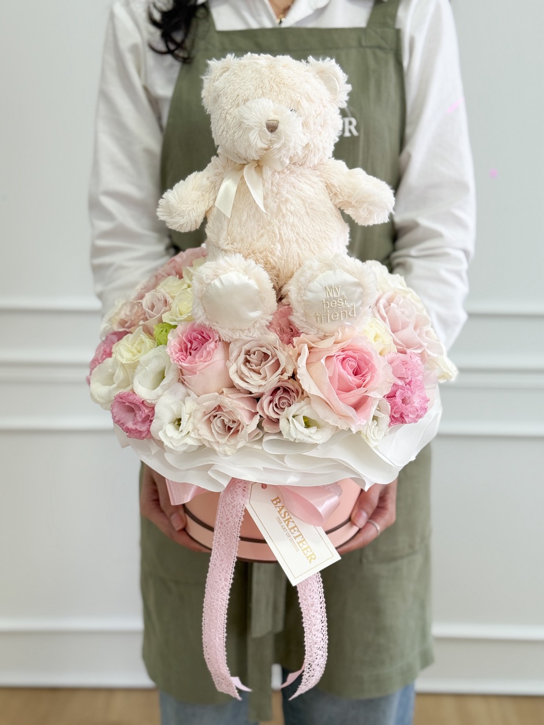 A person in a green apron holds the Pastel Rose & Teddy Celebration Box—a round arrangement of pink and white roses topped with a cream-coloured teddy bear, accented by a pink ribbon and a 