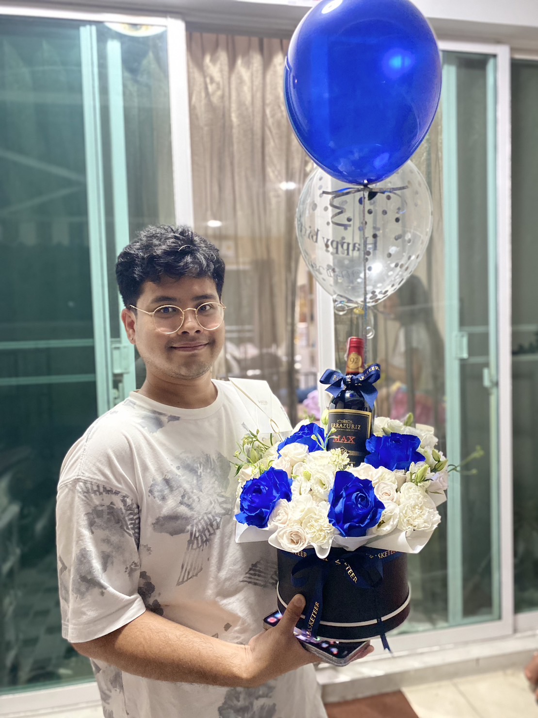 A person with glasses and curly hair is holding a decorative arrangement of blue and white flowers, a bottle of Johnnie Walker Black Label whisky, and a clear balloon with a blue balloon inside. A beautifully wrapped box sits nearby, in front of a window with white curtains.