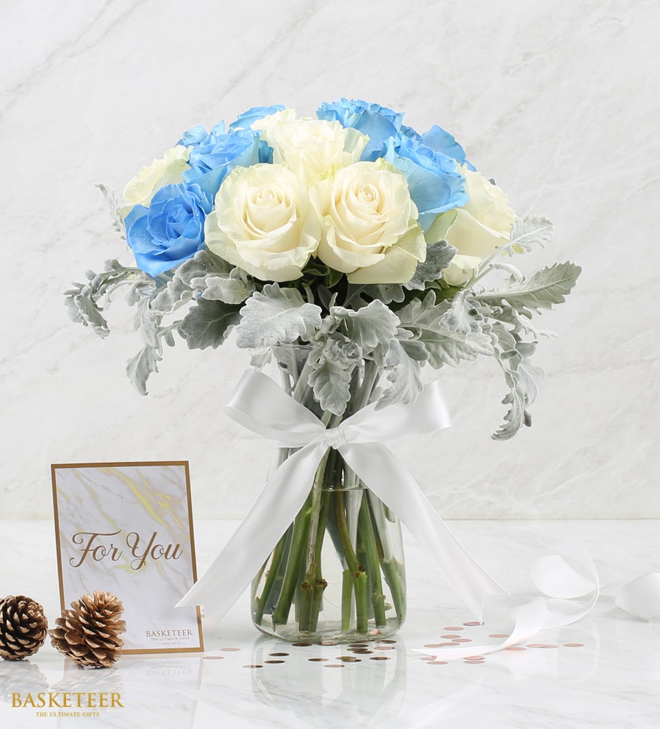 A clear glass vase elegantly showcases a luxurious bouquet of pristine white and enchanting blue roses, delicately accented with silvery-green foliage. The arrangement is tastefully tied with an exquisite white ribbon, bearing the distinguished Basketeer logo. Nearby, charming pine cones and scattered decorative coins add a touch of rustic sophistication. A heartfelt card inscribed with 