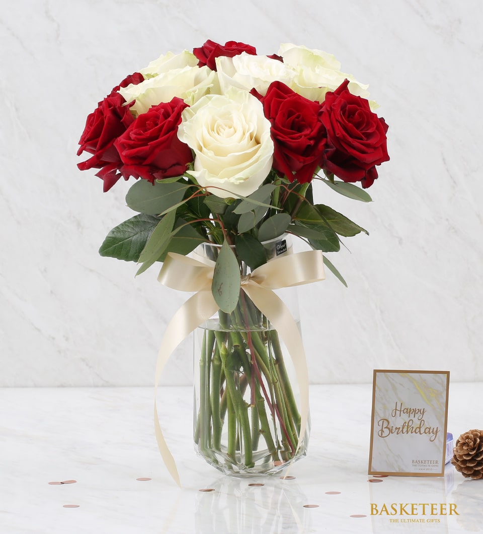A luxurious bouquet of opulent red and pure white roses elegantly arranged in a clear glass vase adorned with a chic white ribbon. This stunning arrangement graces a pristine white marble surface. A thoughtfully crafted small card inscribed with 
