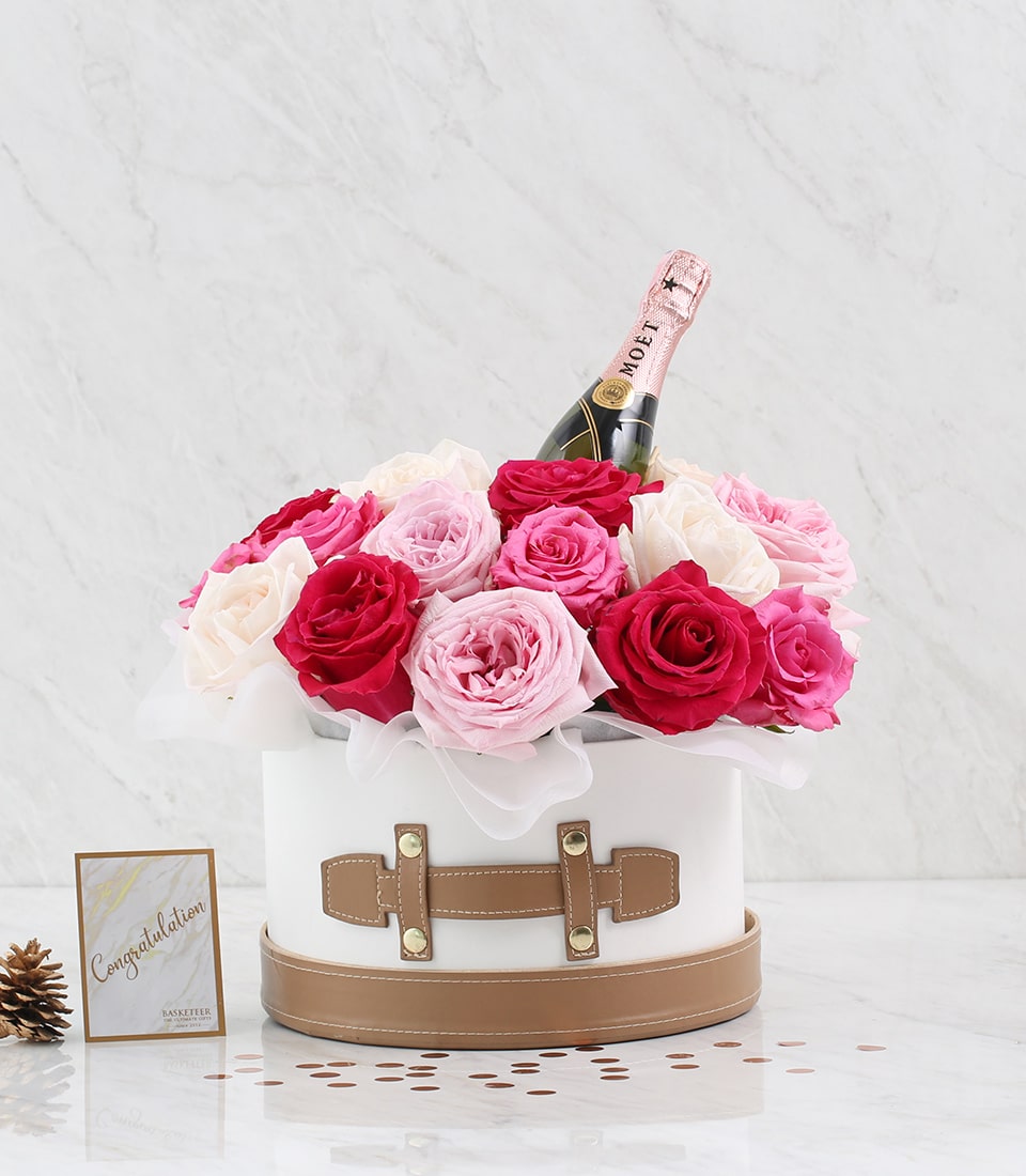 A luxurious white and beige Basketeer box brimming with exquisite pink and red roses encircles a chic Moët Mini, all perfectly arranged on an elegant marble surface. A graceful 
