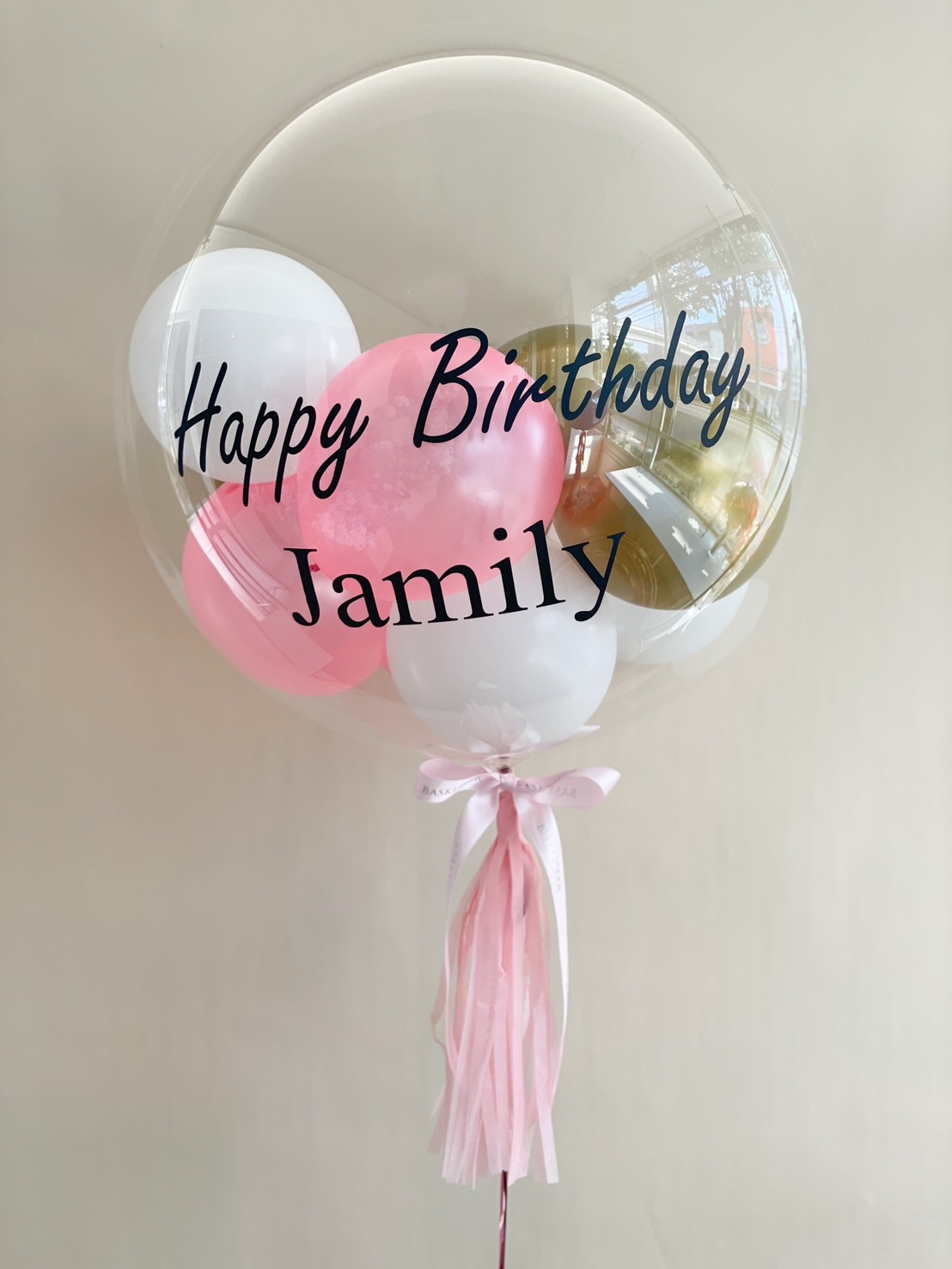 Elegant Happy Birthday bubble balloon with a personalized name, soft pink and white mini balloons inside, and a stylish tassel tail. Perfect for birthday surprises!
