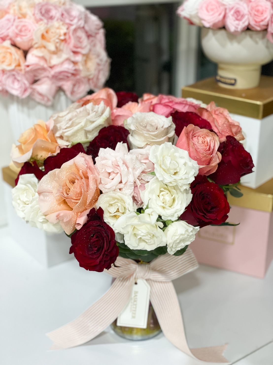 A luxurious vase arrangement of red, white, peach, and blush roses, elegantly tied with a soft pink ribbon.