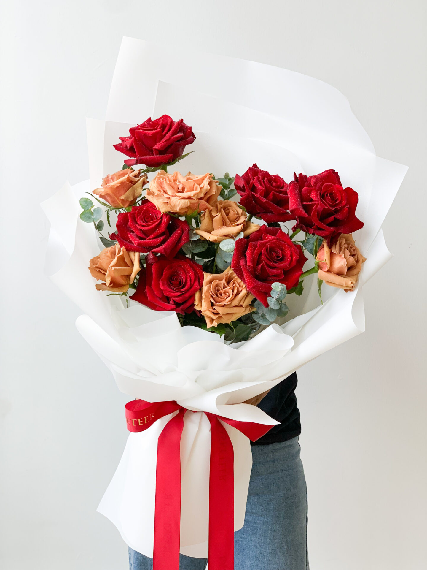 A person is holding a bouquet of red and peach-colored roses wrapped in white paper, reminiscent of a classic Wine & Red Flower Combo. The bouquet is tied with a red ribbon, with the plain white background drawing focus to the vibrant and contrasting colors of the roses.