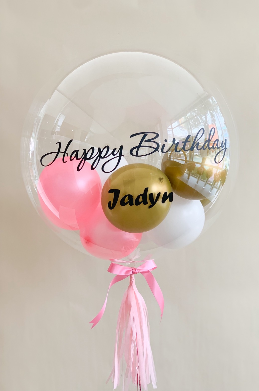 Transparent birthday bubble balloon with personalised name, filled with pink, gold, and white balloons