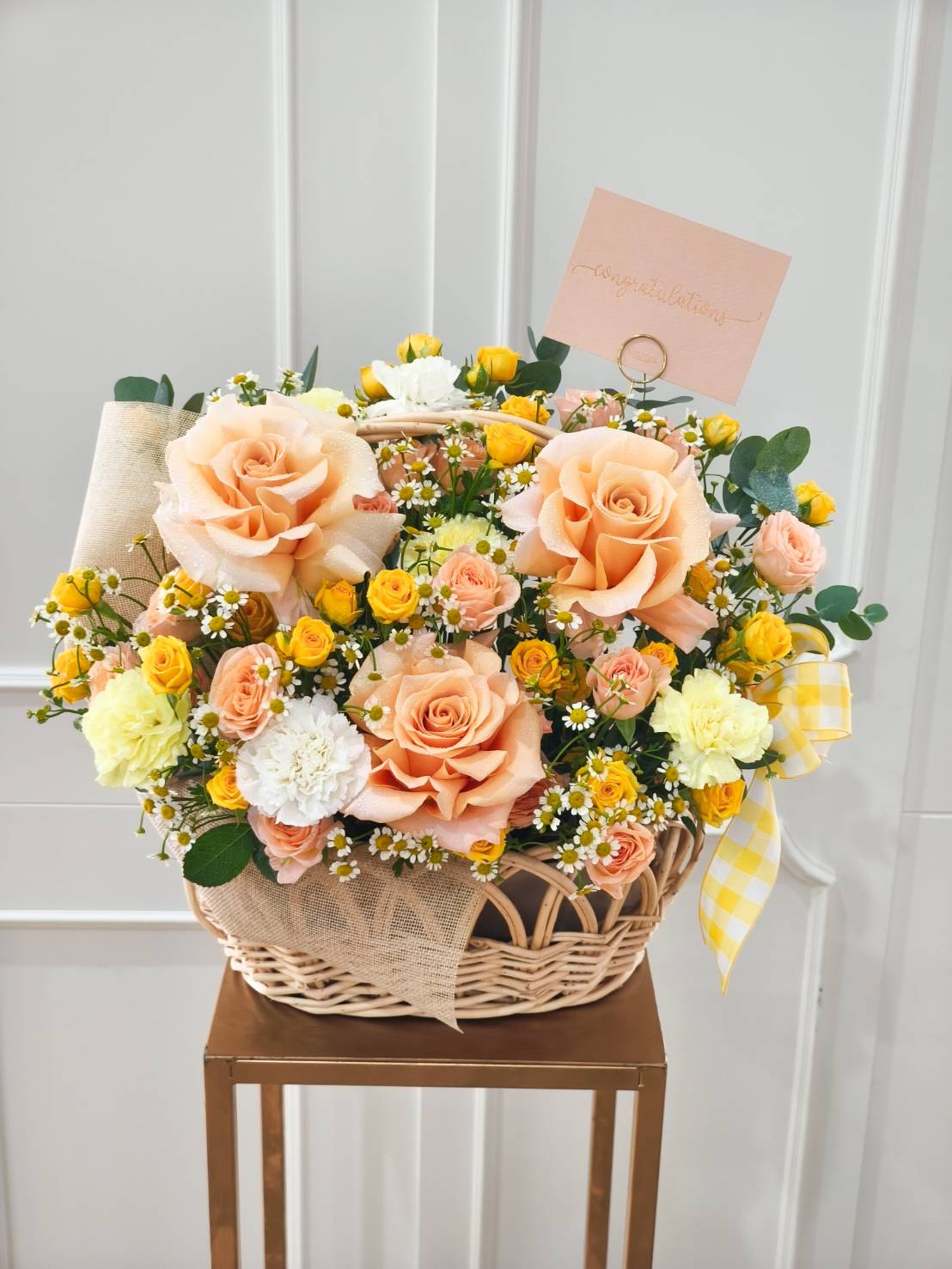 Cheerful basket arrangement with peach roses, yellow spray roses, carnations, and chamomile flowers, decorated with greenery and a gingham ribbon.
