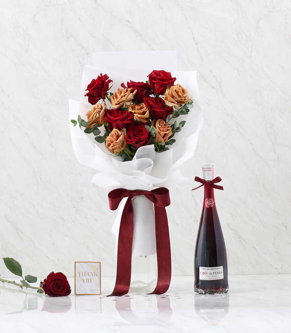 A bouquet of red and peach roses wrapped in white paper with a deep red ribbon sits in a clear vase. Beside it, the elegant Wine & Red Flower Combo includes a 
