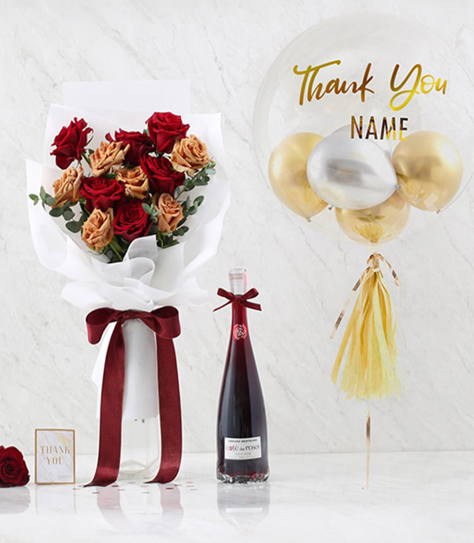 A luxurious thank-you gift set featuring a bouquet of red and peach roses tied with a burgundy ribbon, a bottle of fine wine, and a personalized transparent balloon with gold accents.