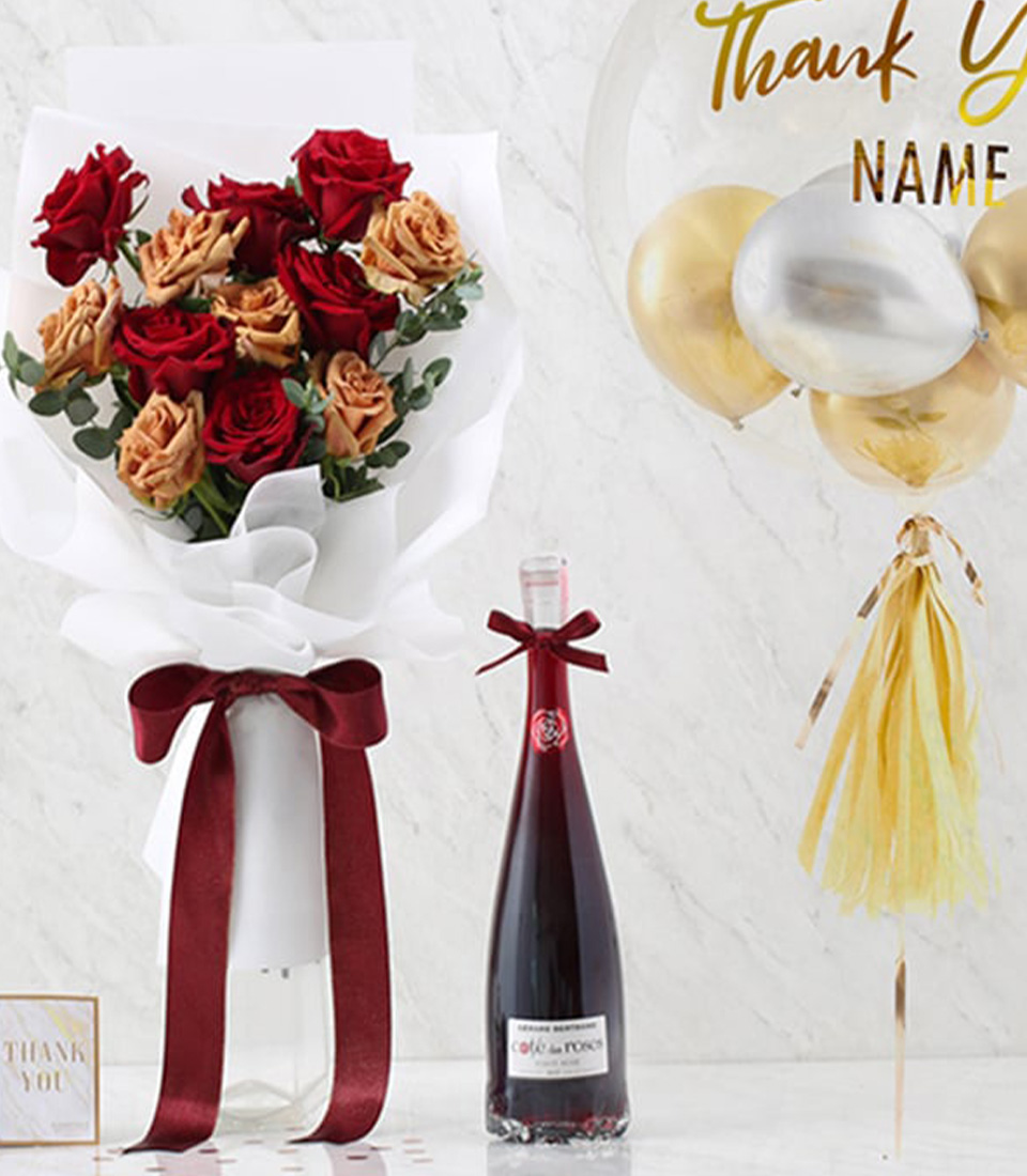 A luxurious thank-you gift set featuring a bouquet of red and peach roses tied with a burgundy ribbon, a bottle of fine wine, and a personalized transparent balloon with gold accents.