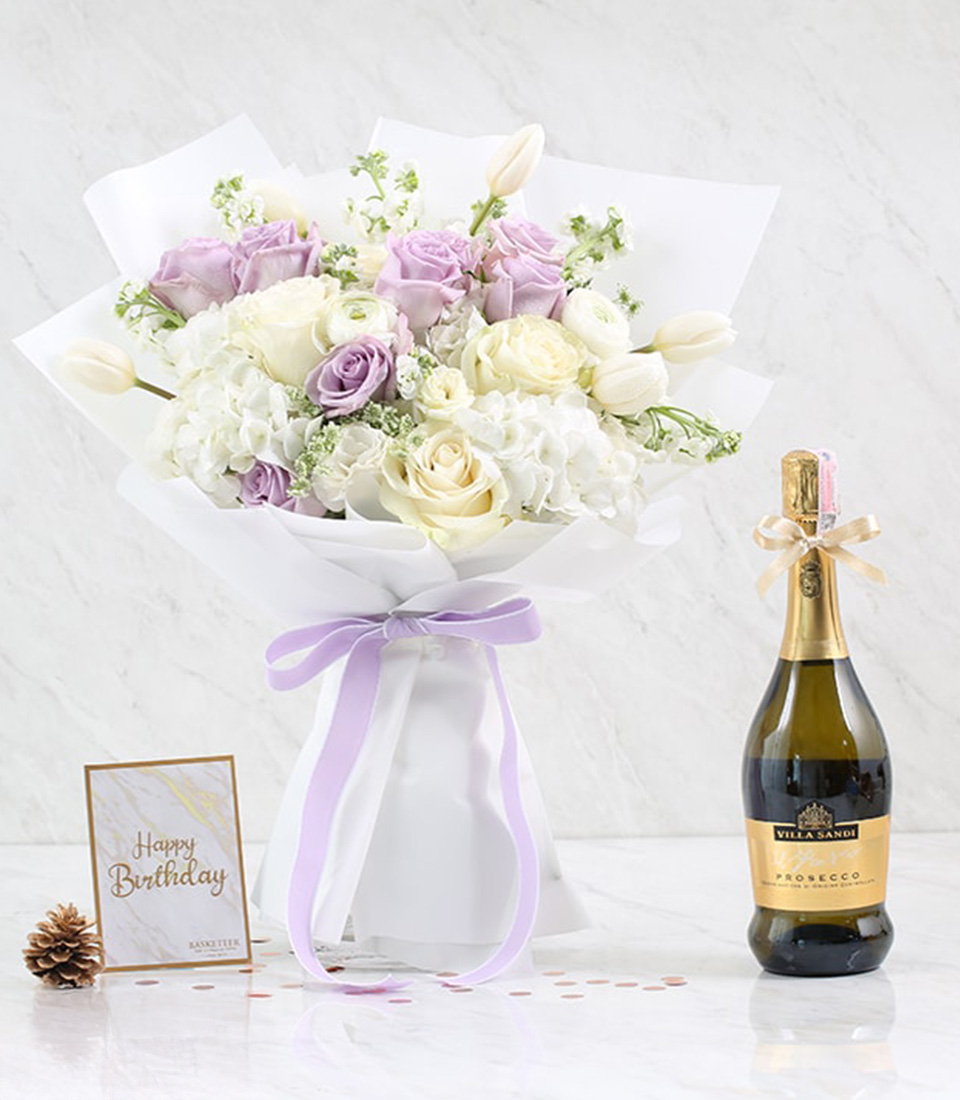 A stunning bouquet of white, lavender, and pale pink roses ensconced in luxurious white wrapping paper with a delicate lavender ribbon bearing the Basketeer logo graces the marble backdrop. Nestled beside this fragrant floral arrangement is a bottle of fine prosecco, exuding elegance and celebration. A lavish gift card inscribed with 