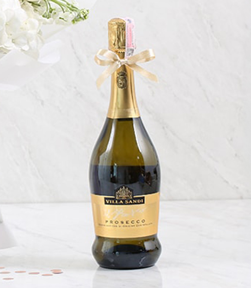 A stunning bouquet of white, lavender, and pale pink roses ensconced in luxurious white wrapping paper with a delicate lavender ribbon bearing the Basketeer logo graces the marble backdrop. Nestled beside this fragrant floral arrangement is a bottle of fine prosecco, exuding elegance and celebration. A lavish gift card inscribed with 